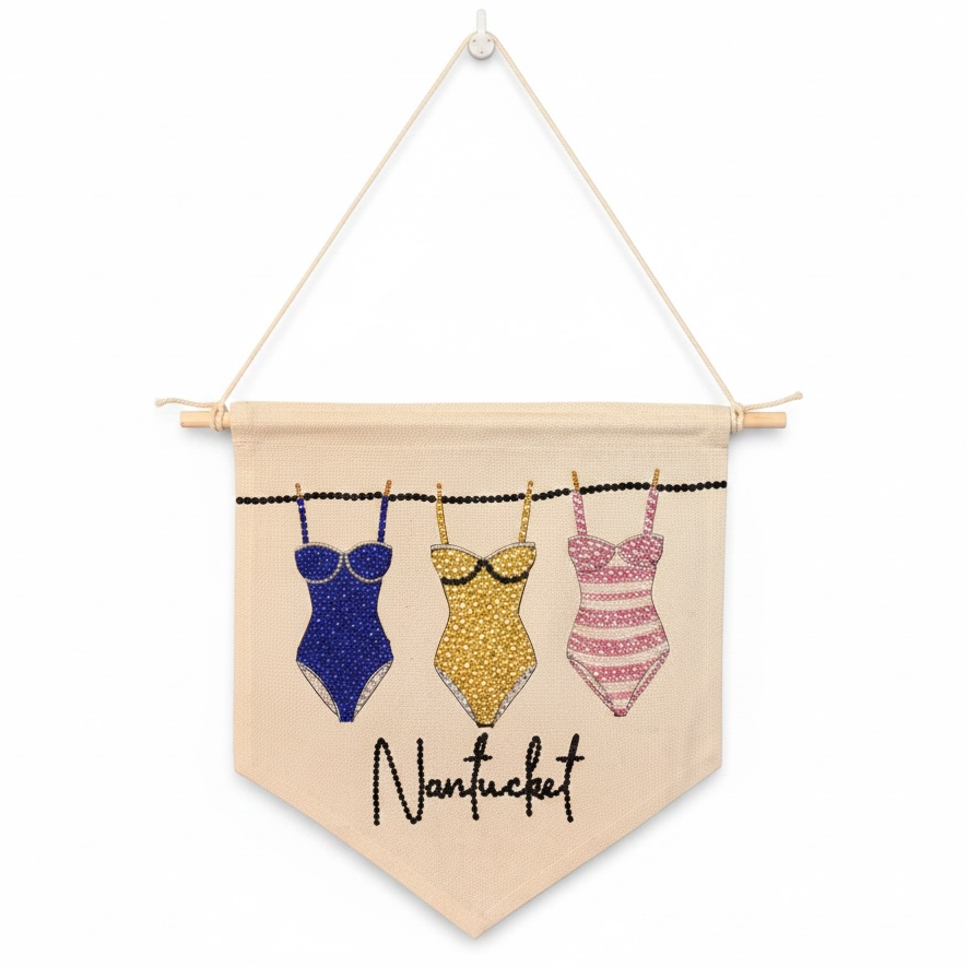 A triangular fabric wall banner with a wooden dowel and hanging cord, featuring three glittery swimsuit designs in blue, yellow, and pink, with the word 'Nantucket' written in black script below.
